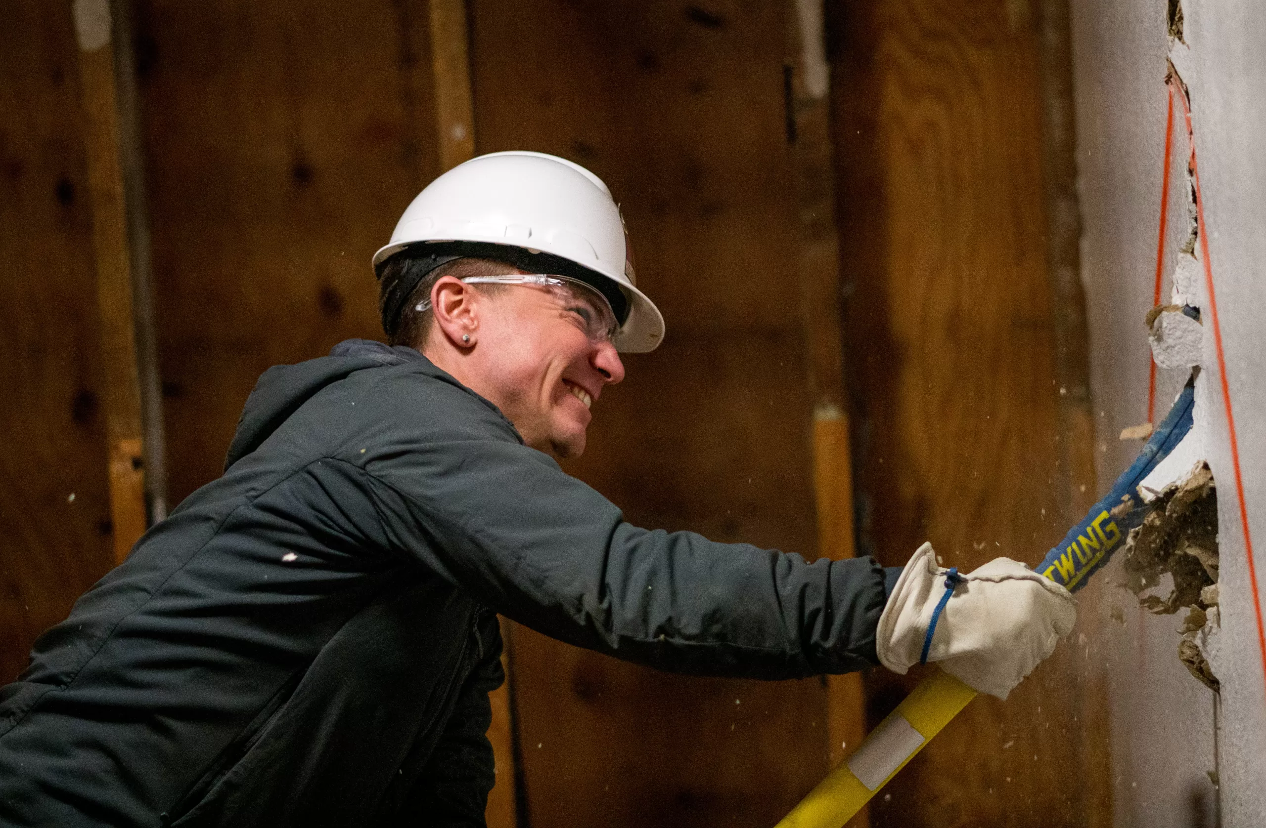 man wearing construction helmet breaks drywall with sledge hammer.