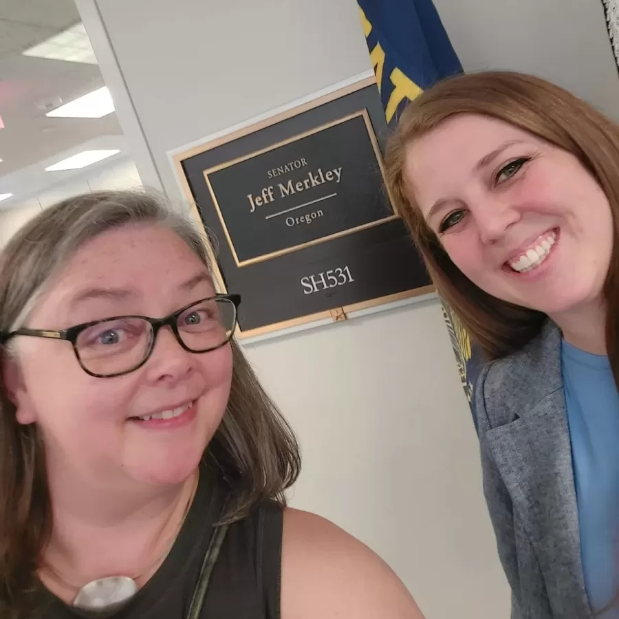 two woman pose in front of a plaque that reads "Senator Jeff Merkley"