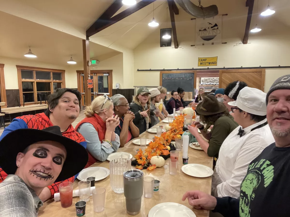 A large group in halloween costumes sits at a table preparing for a meal.